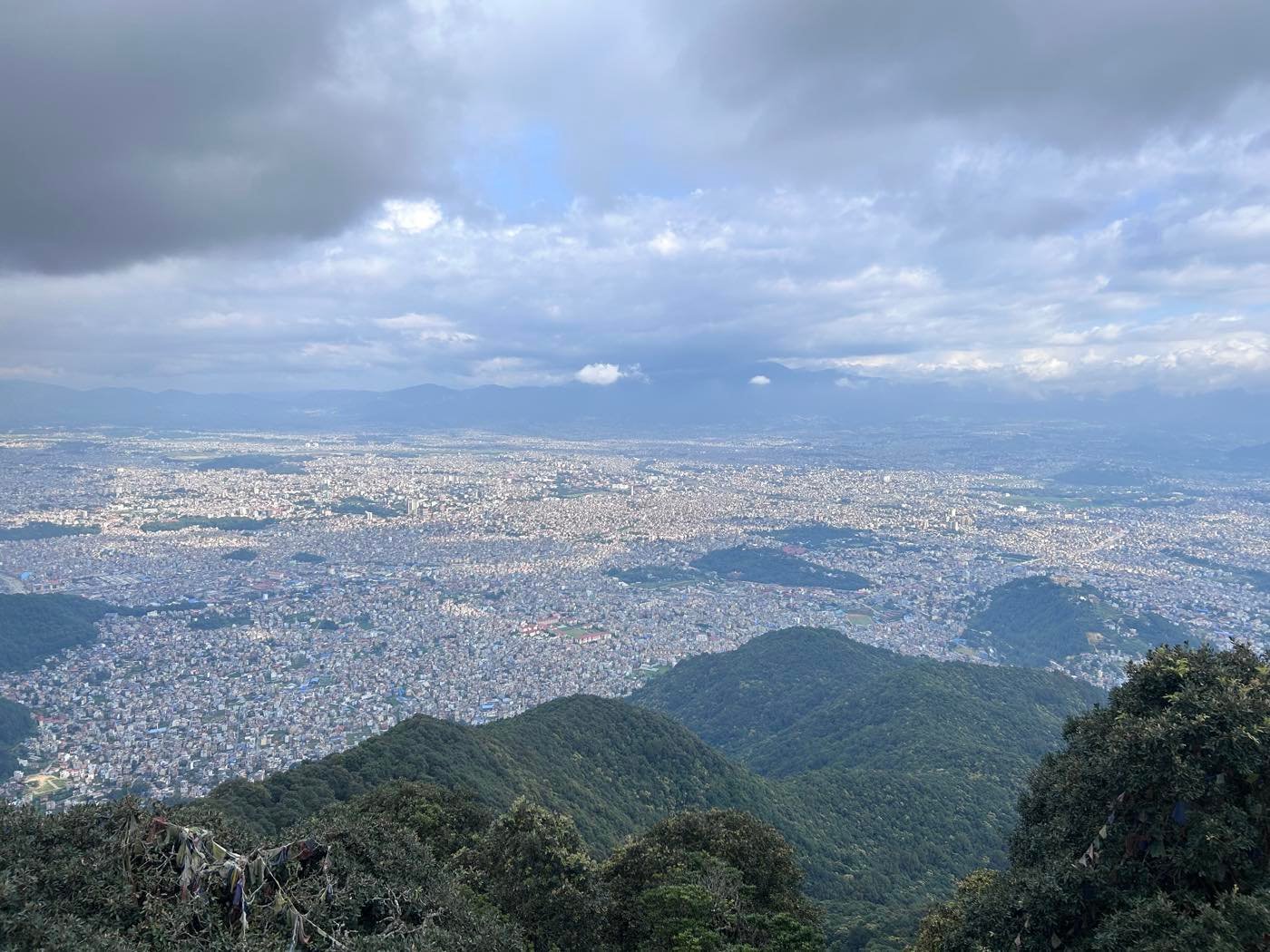 Panoramic view over the Kathmandu Valley and its sprawling cityscape from a forested hilltop, prayer flags in the foreground
